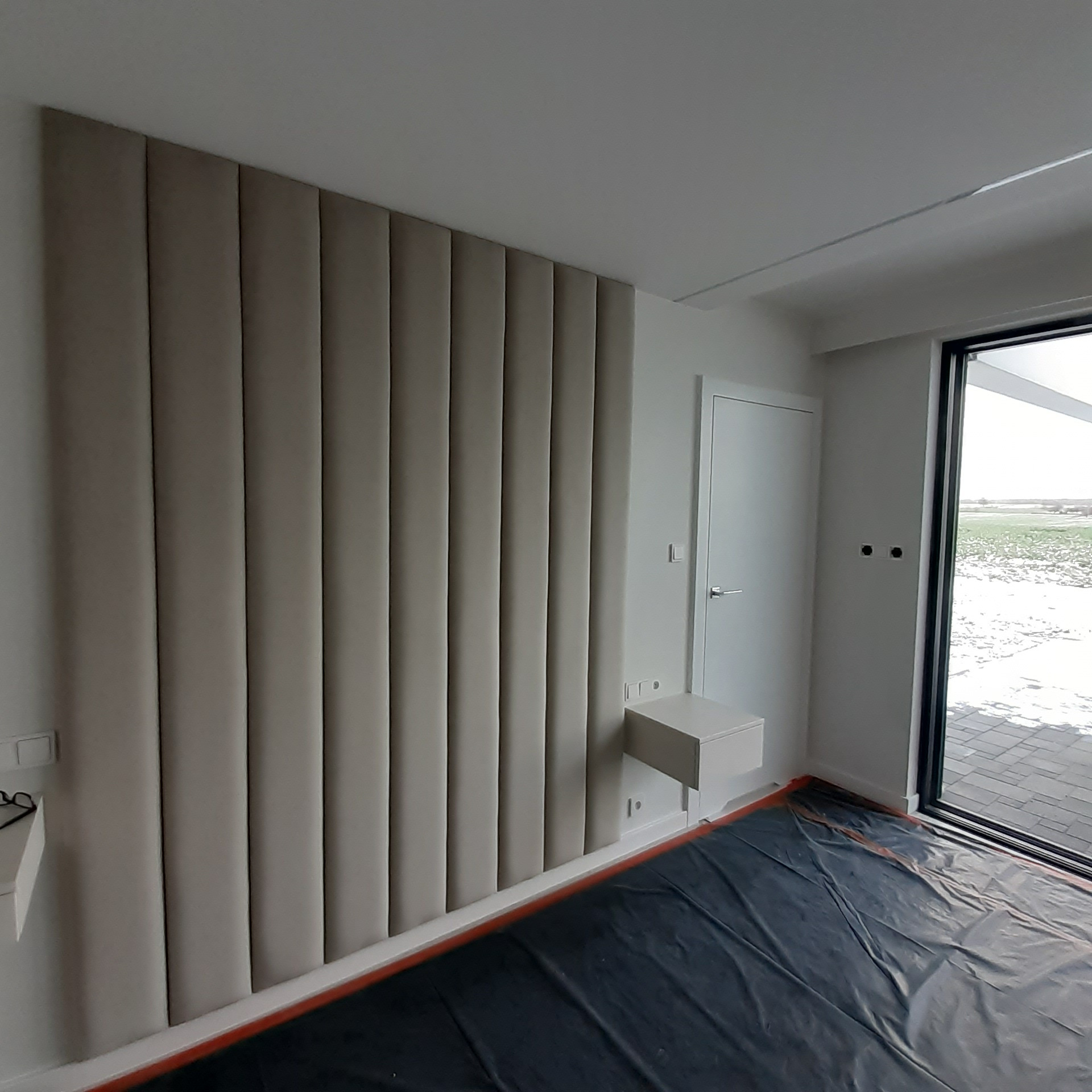 Beżowy panel tapicerowany pionowymi pasami na ścianie w sypialni, obok biała szafka nocna i drzwi balkonowe, podłoga zabezpieczona folią malarską.