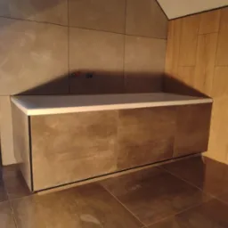 Nowoczesna zabudowa wanny z płytkami imitującymi beton, widoczne przyłącza wodne i fragment okna z widokiem na zewnątrz.