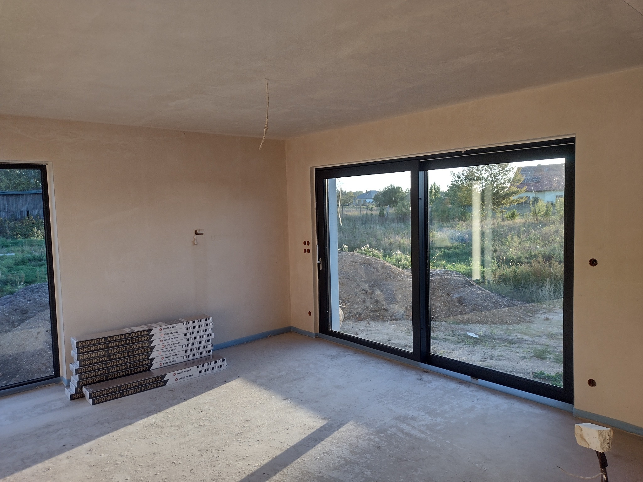 Wnętrze w stanie deweloperskim z przygotowanymi paczkami paneli podłogowych Kronopol Aurum Flooring, widok na ogród przez duże okno przesuwne.