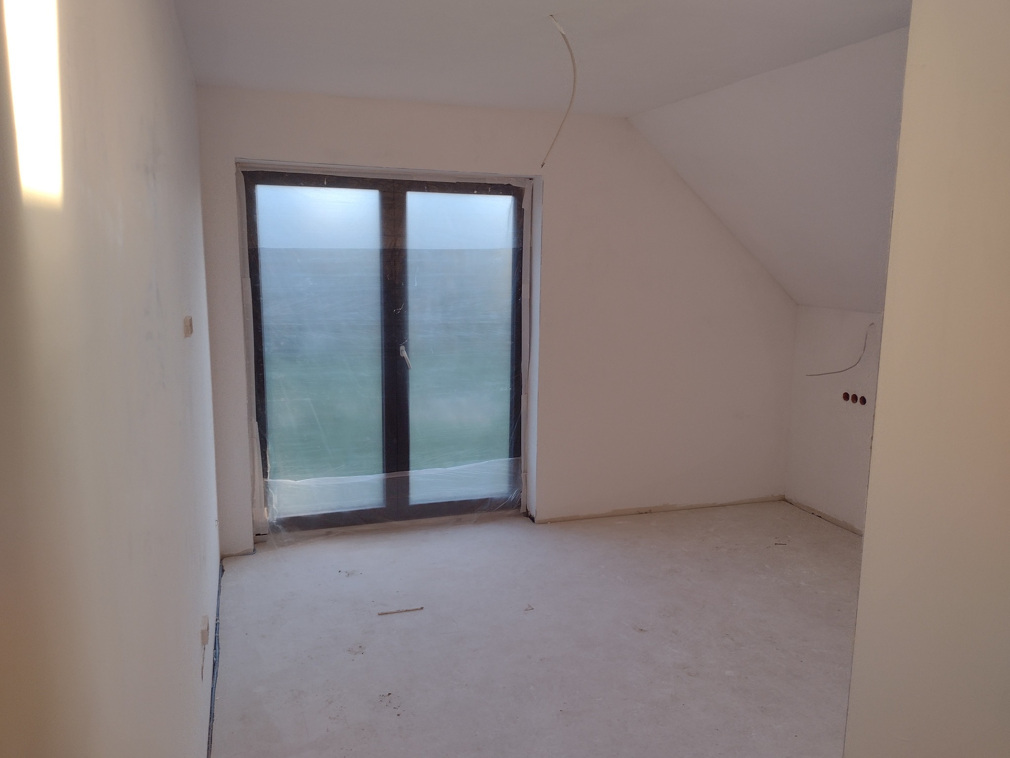 Pusty pokój na poddaszu w trakcie remontu, ściany wyszpachlowane na gładko, okno zabezpieczone folią malarską, widoczne gniazdka elektryczne.