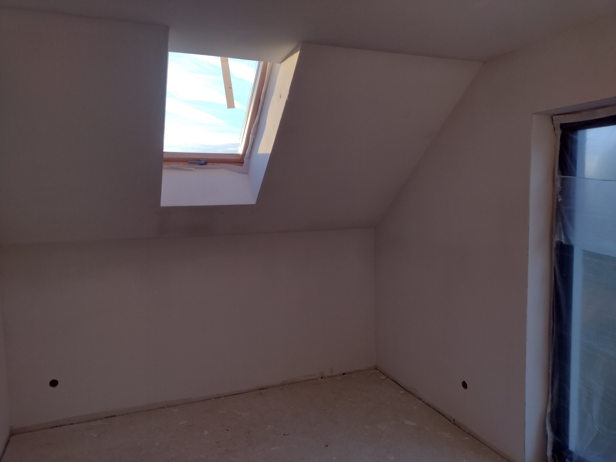 Poddasze po szpachlowaniu, widoczne gładkie ściany, okno dachowe z widokiem na niebo i okno zabezpieczone folią malarską.