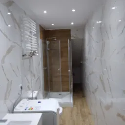 Wnętrze łazienki po remoncie z kabiną prysznicową, umywalką, pralką i grzejnikiem drabinkowym, wykończone białymi płytkami imitującymi marmur i drewnopodobną podłogą.