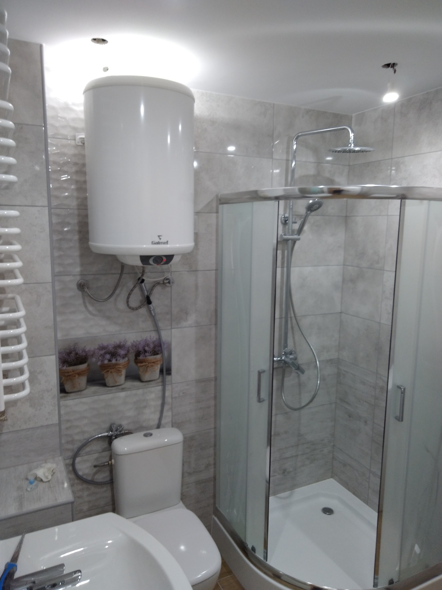 Nowoczesna łazienka z kabiną prysznicową, podgrzewaczem wody Galmet, umywalką i toaletą, wykończona płytkami w odcieniach szarości.