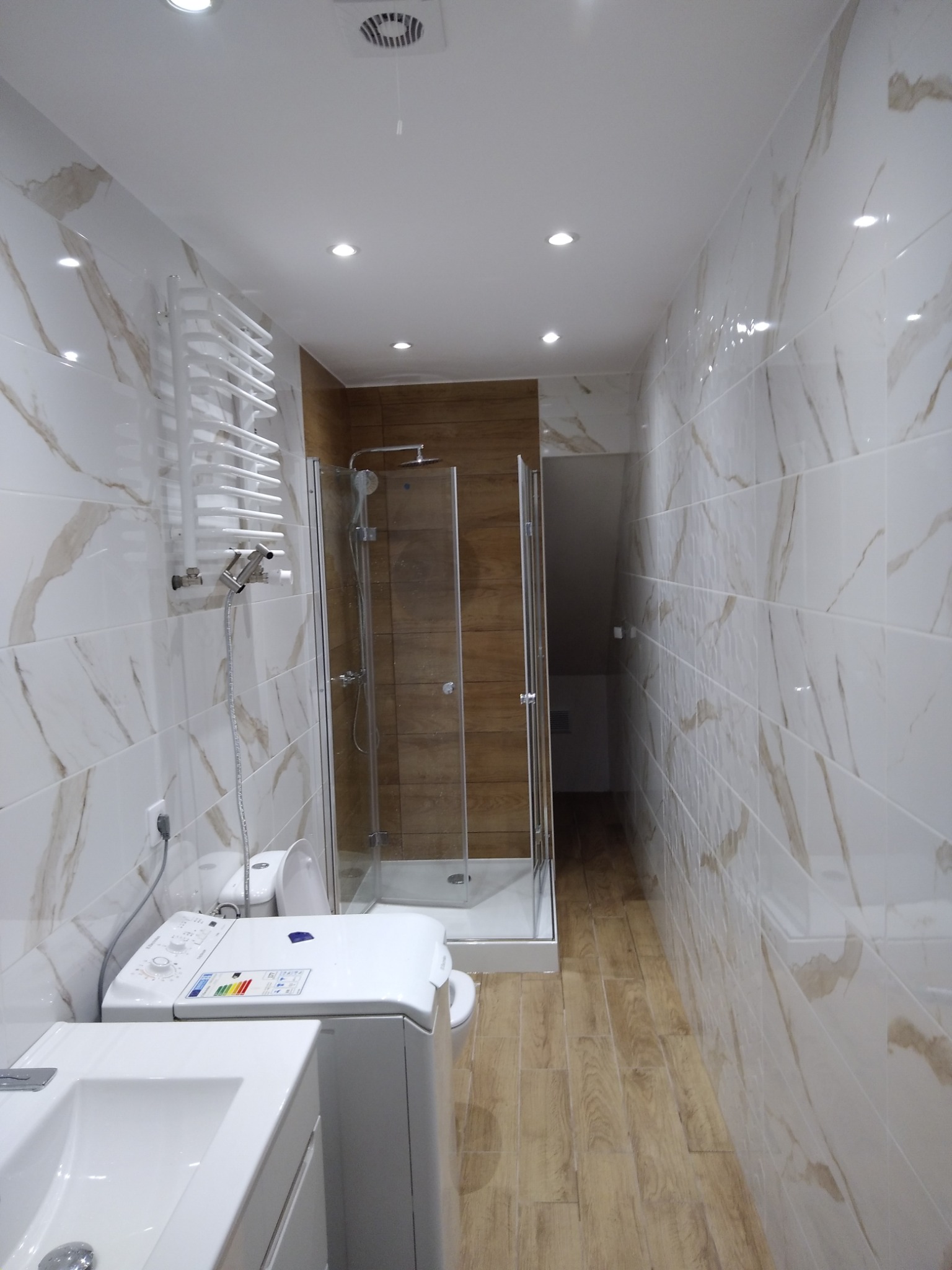 Wnętrze łazienki po remoncie z kabiną prysznicową, umywalką, pralką i grzejnikiem drabinkowym, wykończone białymi płytkami imitującymi marmur i drewnopodobną podłogą.