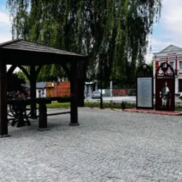 Drewniana altana z kwiatami w donicach na placu wyłożonym kostką brukową, w tle drzewa i budynki.