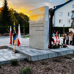 Uroczyste odsłonięcie pomnika z szarego granitu, otoczonego kostką brukową i flagami Polski, w tle zgromadzenie ludzi i budynek mieszkalny.
