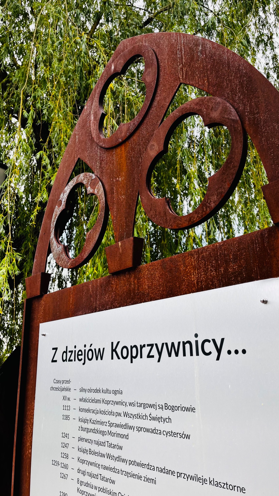 Rdzewiejąca, ażurowa konstrukcja z motywami serca nad tablicą informacyjną z napisem 'Z dziejów Koprzywnicy...', na tle zielonych gałęzi drzewa.