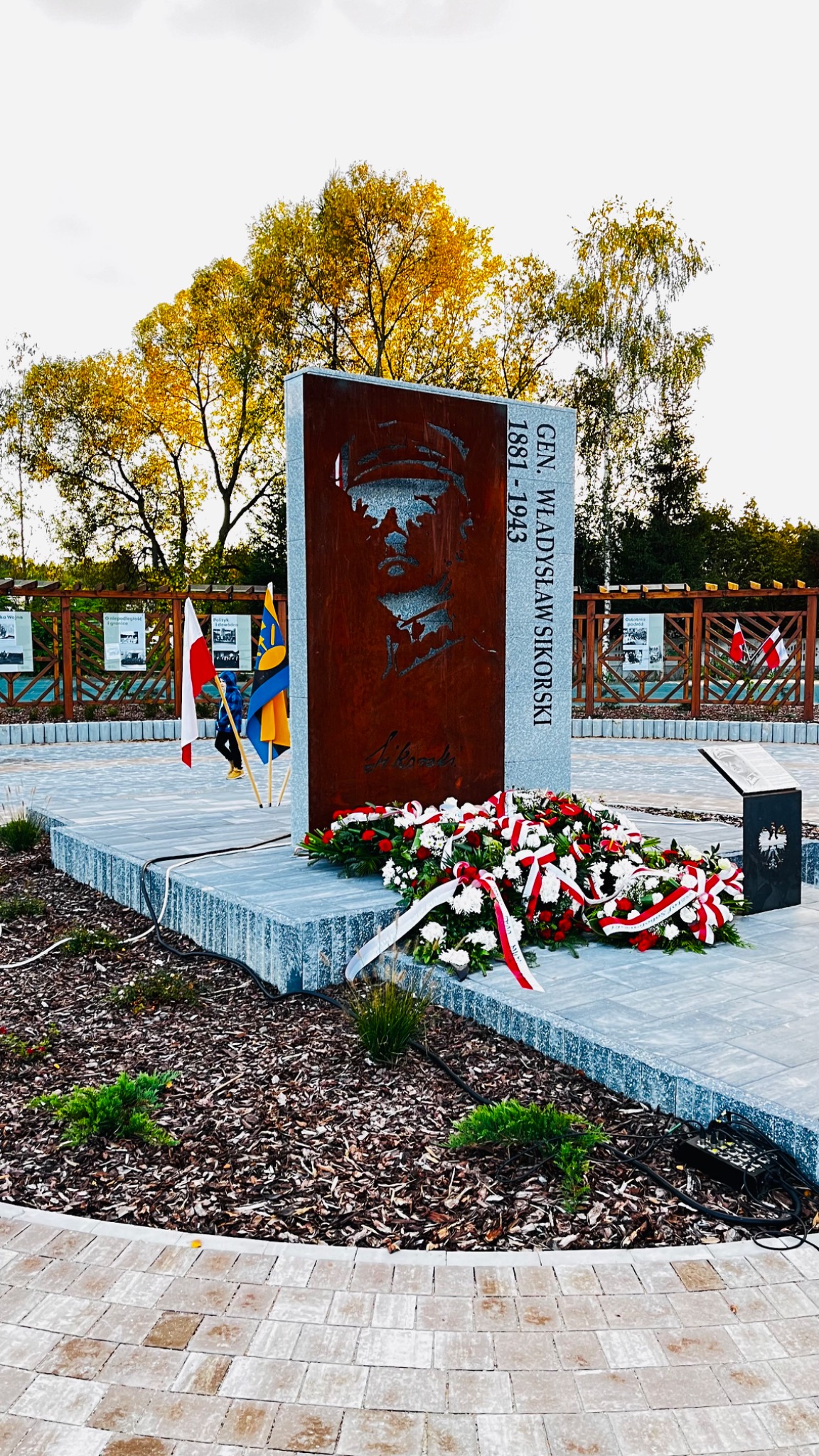 Pomnik generała Władysława Sikorskiego z wyciętym laserowo portretem na rdzawym tle, otoczony wieńcami z białych i czerwonych kwiatów, z flagami Polski i Ukrainy w tle, na placu wyłożonym kostką...