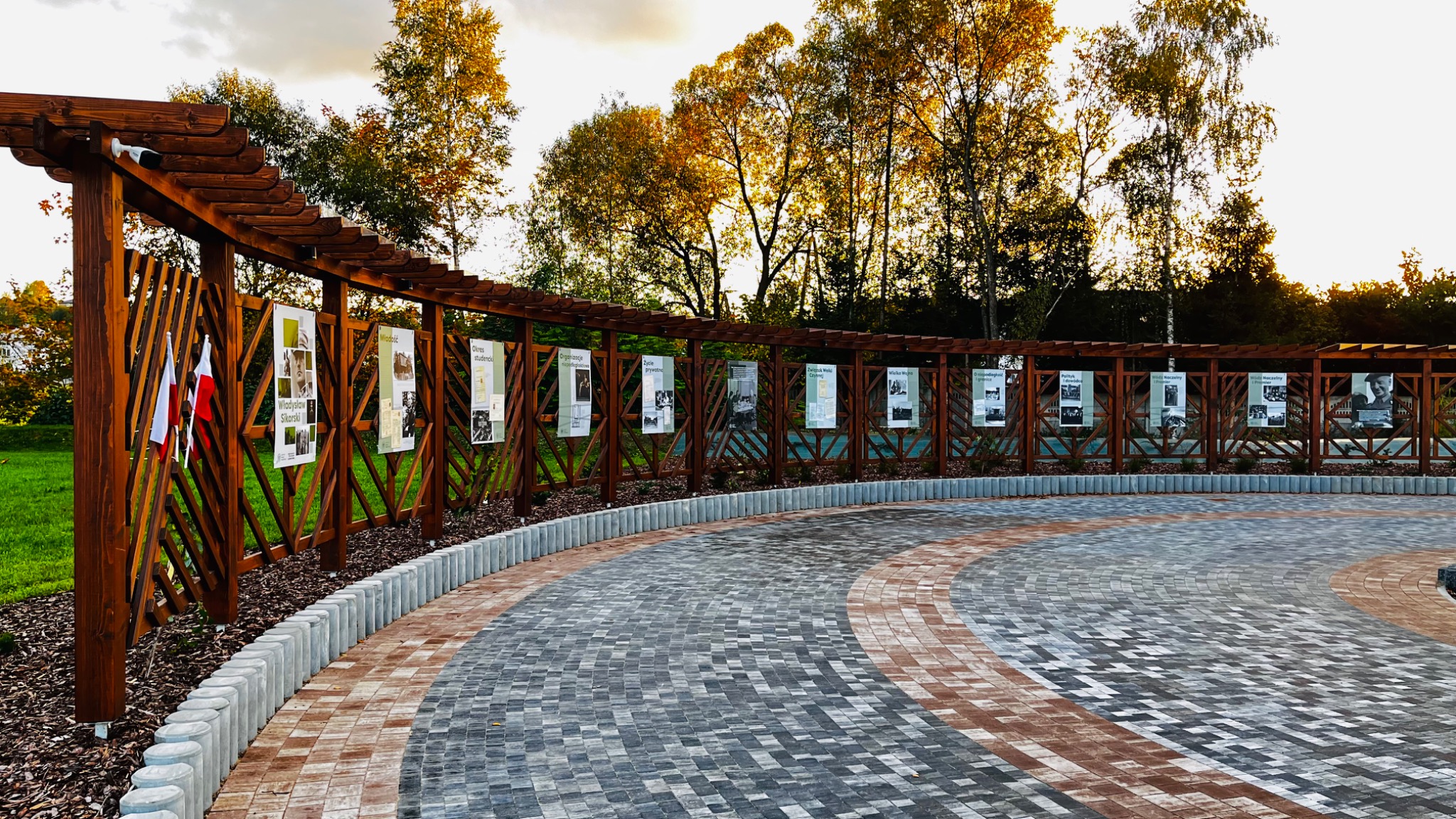 Oświetlona drewniana pergola z ekspozycją plakatów historycznych i polskimi flagami, otoczona brukowaną ścieżką w parku.