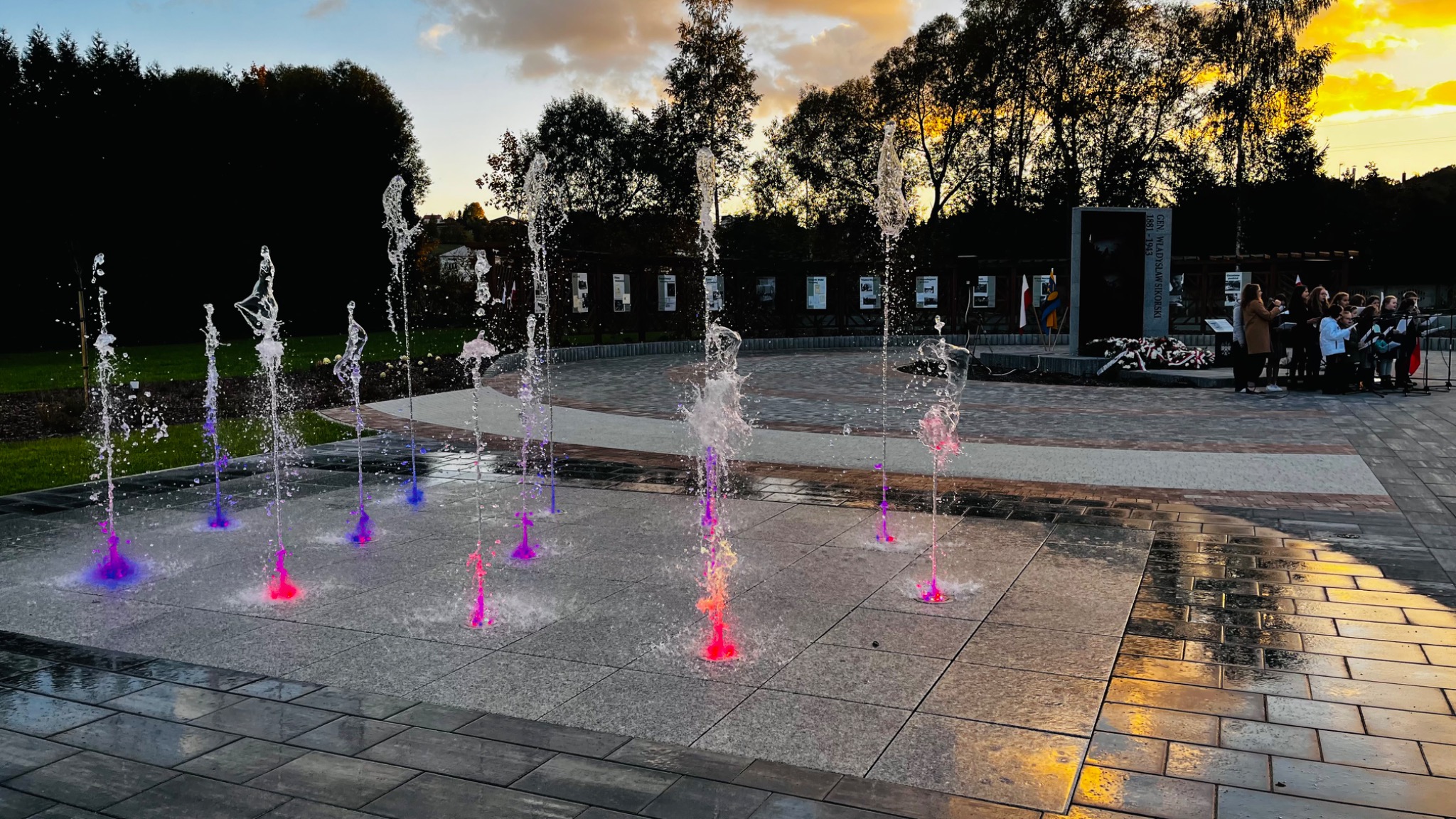 Podświetlana fontanna posadzkowa z kolorowymi strumieniami wody na tle pomnika i grupy osób podczas uroczystości.
