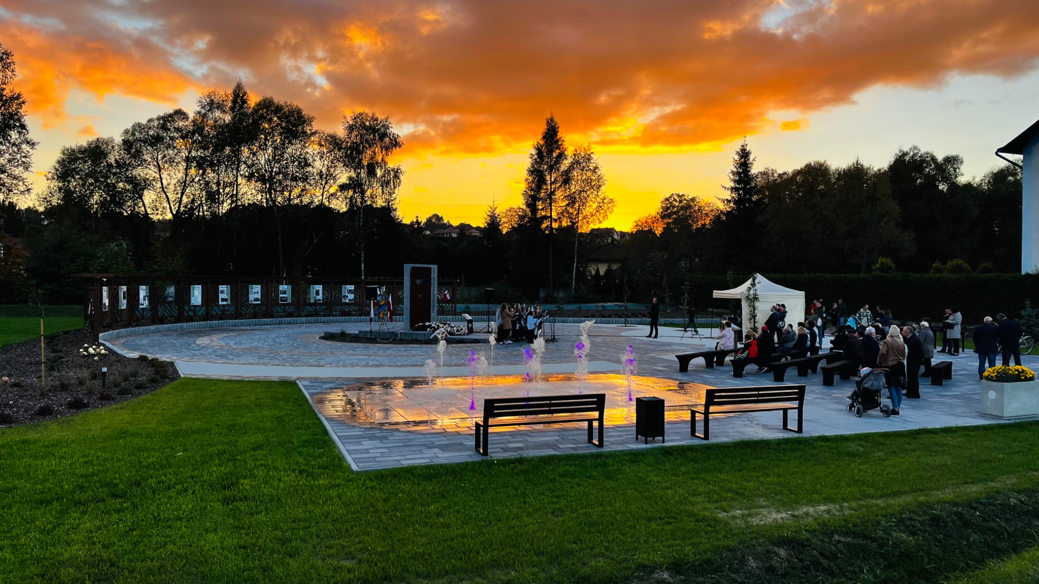 Uroczyste otwarcie placu z podświetlaną fontanną i miejscami do siedzenia, na tle pomnika i zachodzącego słońca.