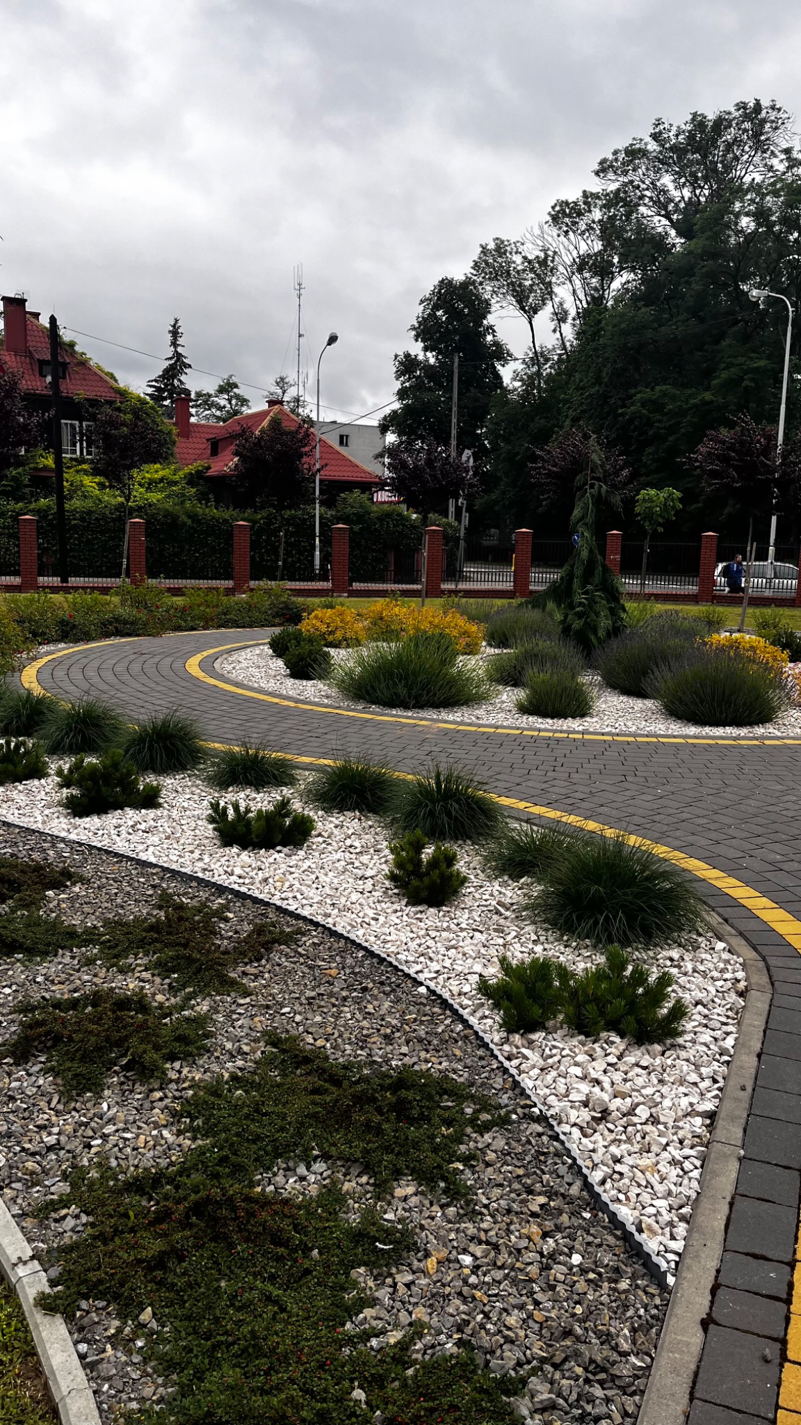 Kręty chodnik z szarej kostki brukowej z żółtymi obwódkami, otoczony białym i szarym żwirem oraz różnorodną zielenią, w tle domy z czerwonymi dachami.