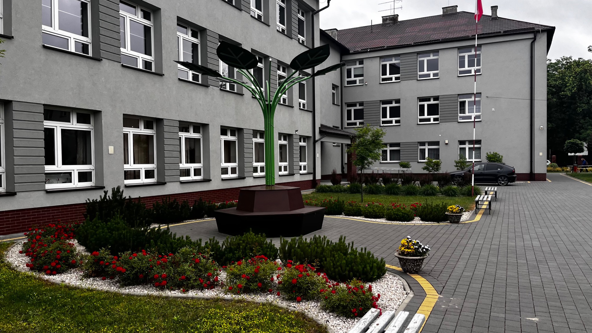 Nowoczesna aranżacja przestrzeni przed budynkiem: metalowa rzeźba drzewa z liśćmi, otoczona rabatami z czerwonymi różami i iglakami, na tle szarego budynku z białymi oknami.