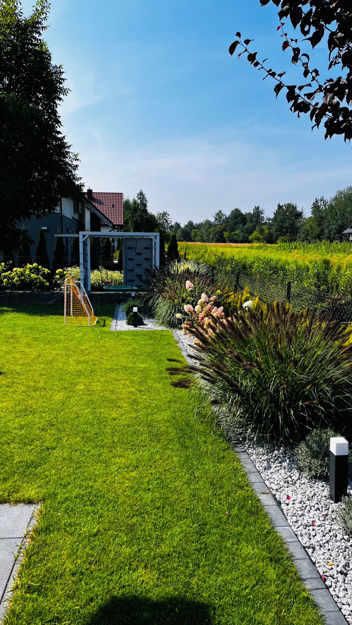 Zadbany ogród przydomowy z równo przystrzyżonym trawnikiem, ozdobnymi trawami, hortensjami, białym żwirem, małą bramką do piłki nożnej i placem zabaw z huśtawką w tle.