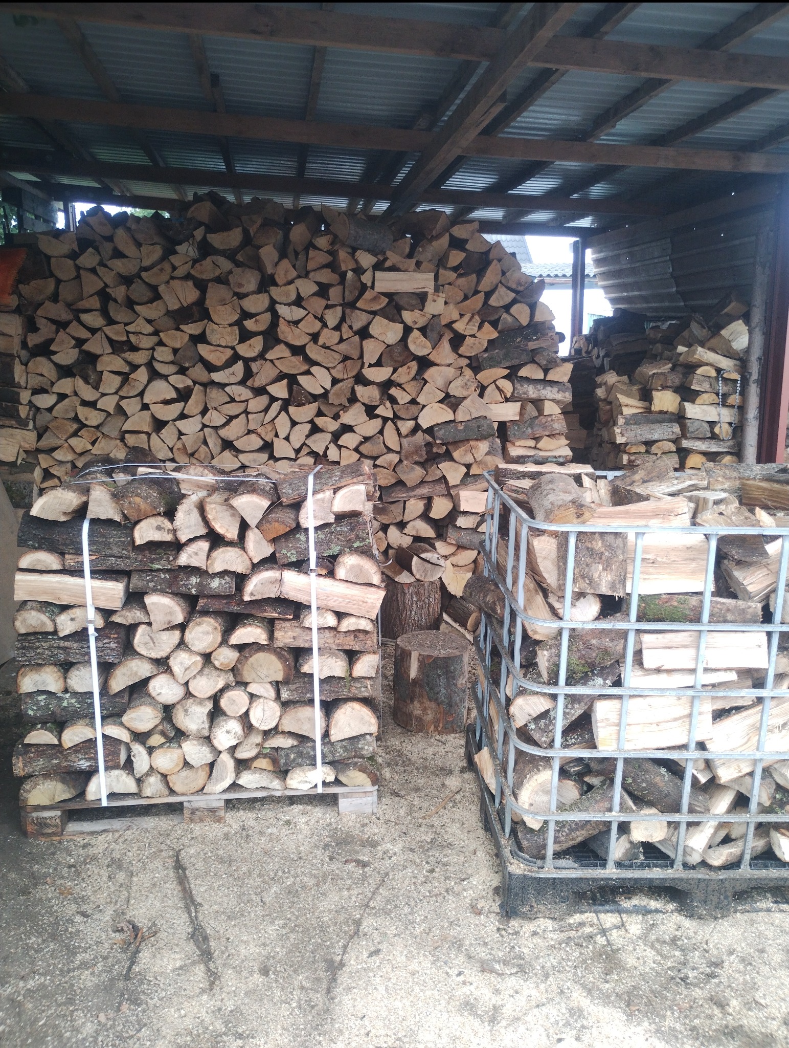 Składowane drewno kominkowe pod zadaszeniem, ułożone w stosy i metalowych koszach. Widoczne kawałki kory i trociny na podłożu. Drewno przygotowane do sezonu grzewczego.
