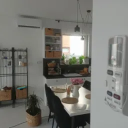 Nowoczesny salon z zamontowanym klimatyzatorem na ścianie, widoczny pilot do klimatyzacji na ścianie, stół jadalny i elementy dekoracyjne.