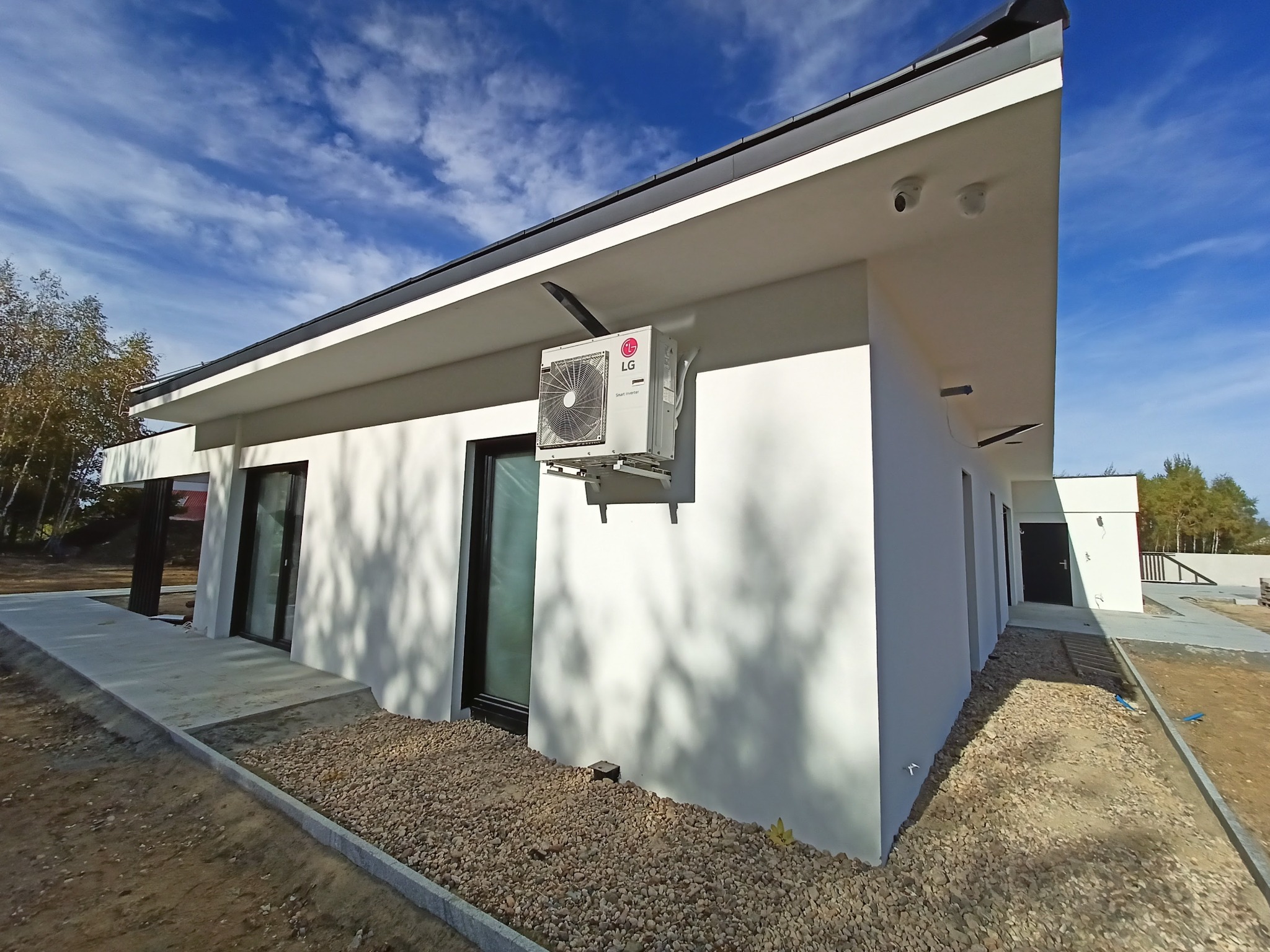 Biały budynek jednorodzinny z zamontowaną jednostką zewnętrzną klimatyzacji LG, widok z boku, błękitne niebo w tle.