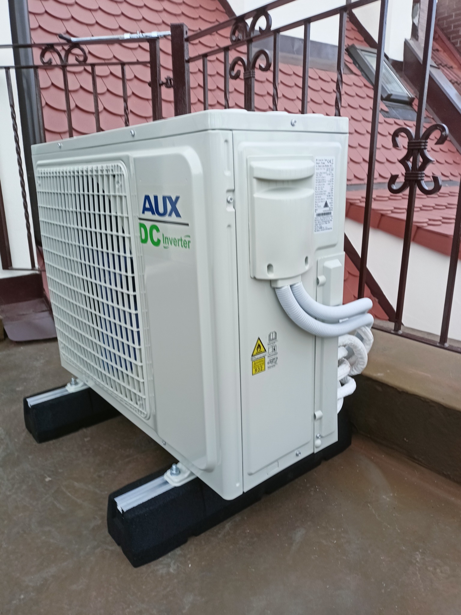 Zewnętrzna jednostka klimatyzacji AUX DC Inverter umieszczona na czarnych, piankowych podstawach, z widocznymi przewodami i balustradą balkonu w tle.