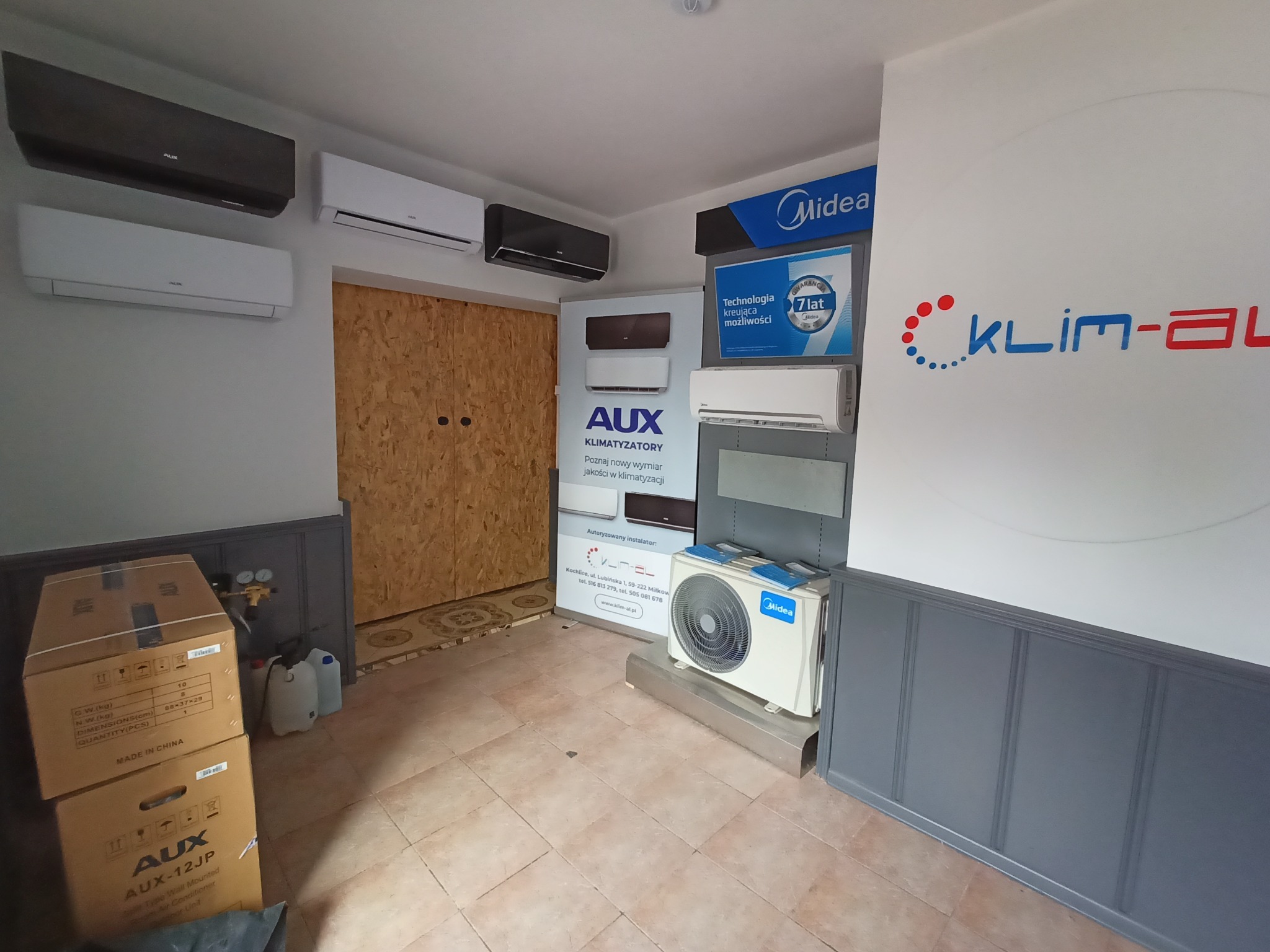 Ekspozycja klimatyzatorów różnych marek i modeli w salonie, widoczne jednostki wewnętrzne AUX i Midea, kartony z nowymi urządzeniami oraz logo firmy Klim-al na ścianie.