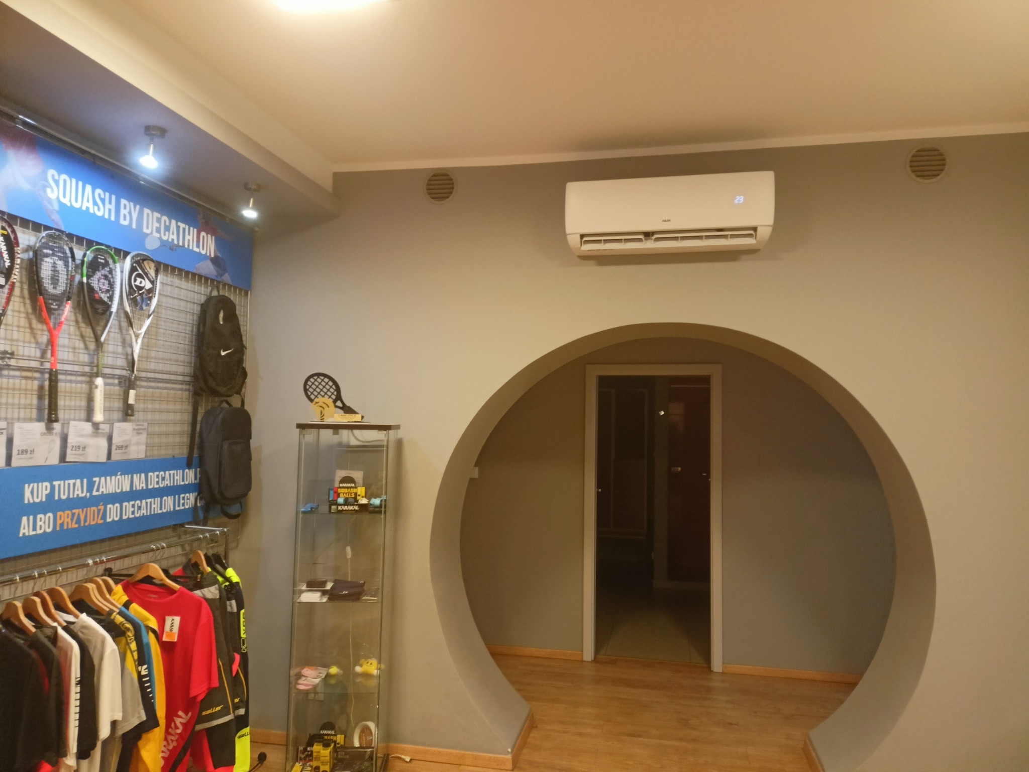 Wnętrze sklepu sportowego z klimatyzatorem AUX na ścianie, widoczne rakiety do squasha, odzież sportowa i okrągłe przejście do innego pomieszczenia.