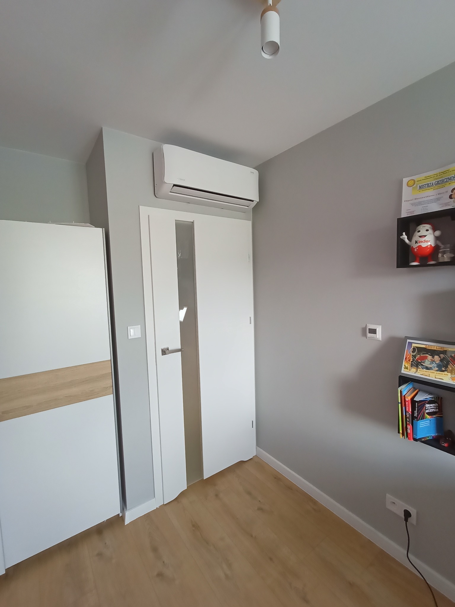 Biały klimatyzator ścienny zamontowany nad drzwiami w pokoju z szarą ścianą, drewnianą podłogą i półką z książkami.