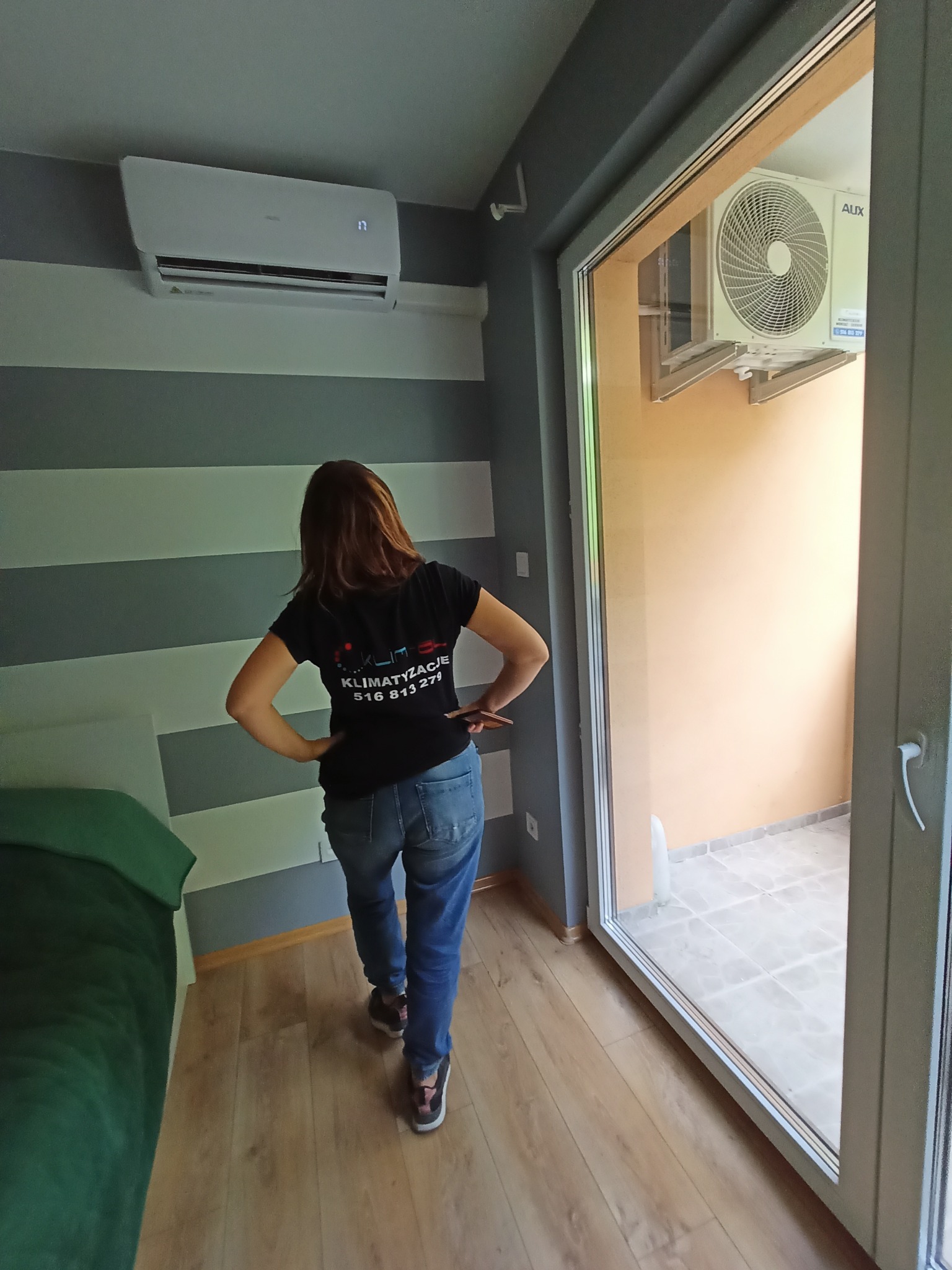 Wnętrze pokoju z zamontowaną klimatyzacją ścienną AUX oraz widok na jednostkę zewnętrzną przez otwarte drzwi balkonowe. Osoba w koszulce z logo firmy klimatyzacyjnej stoi tyłem do kamery.