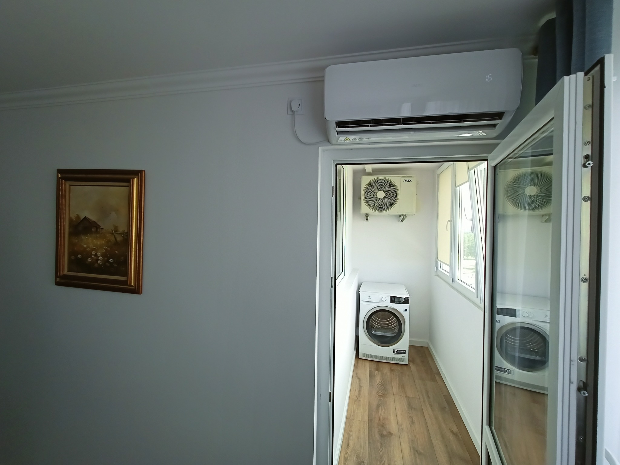 Wnętrze balkonu z zamontowaną klimatyzacją AUX, widoczna jednostka wewnętrzna na ścianie i zewnętrzna na balkonie, obok pralka automatyczna, na ścianie wisi obrazek