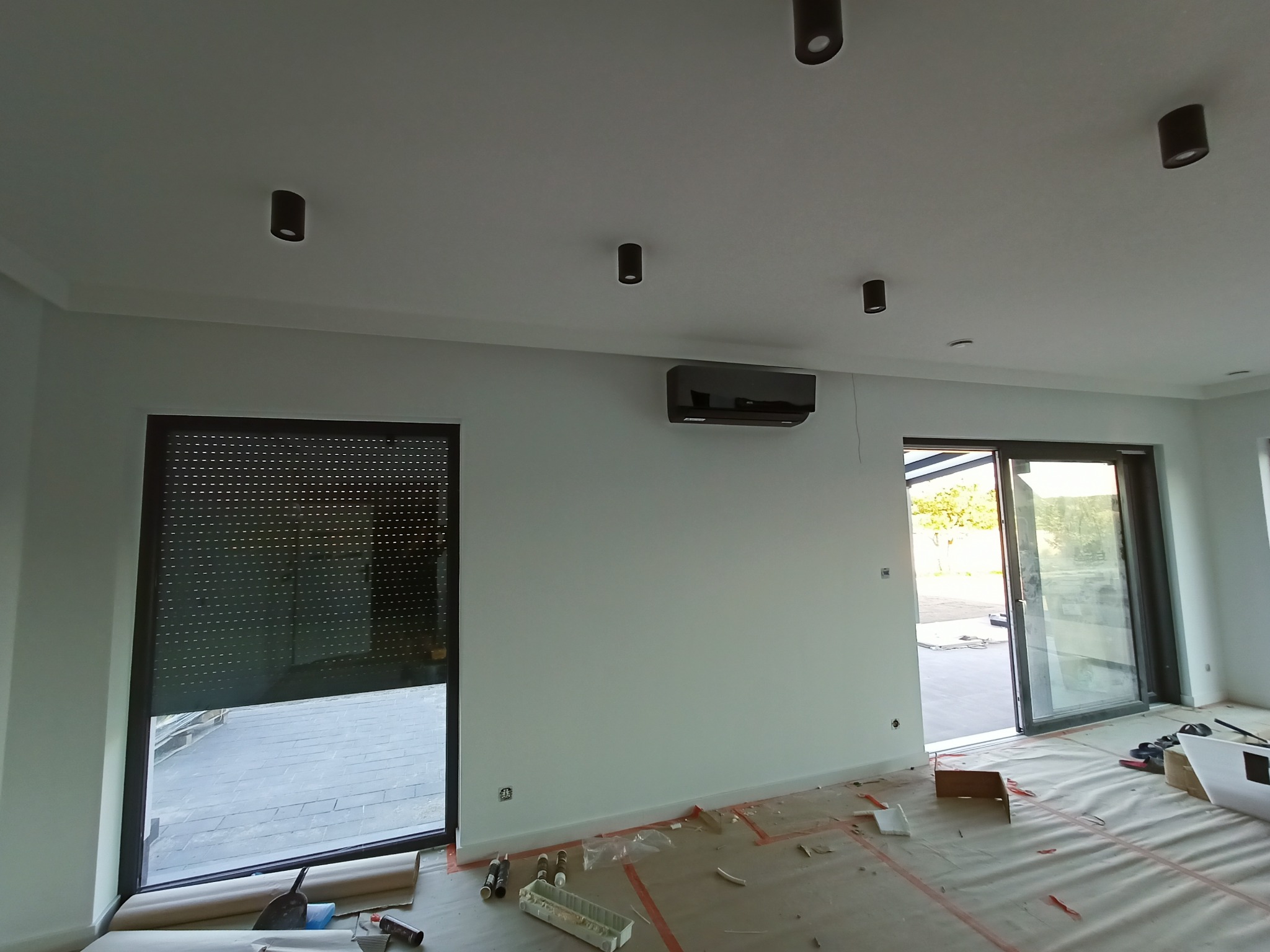 Czarny klimatyzator ścienny zamontowany w jasnym, przestronnym pokoju z dużymi oknami i czarnymi roletami, widoczne prace wykończeniowe, podłoga zabezpieczona folią ochronną.