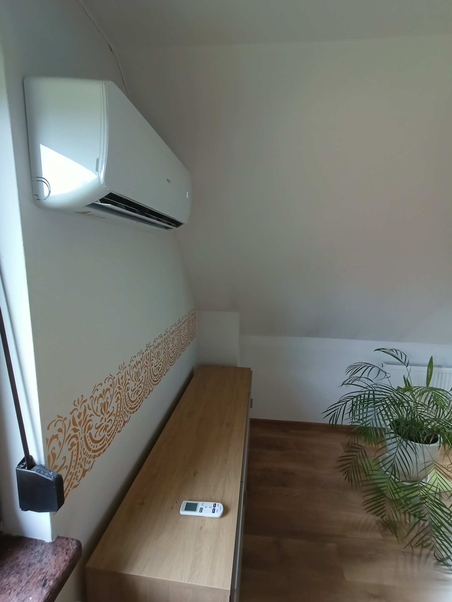 Biały klimatyzator ścienny, zamontowany w pokoju na poddaszu z drewnianą komodą, pilotem i rośliną doniczkową.