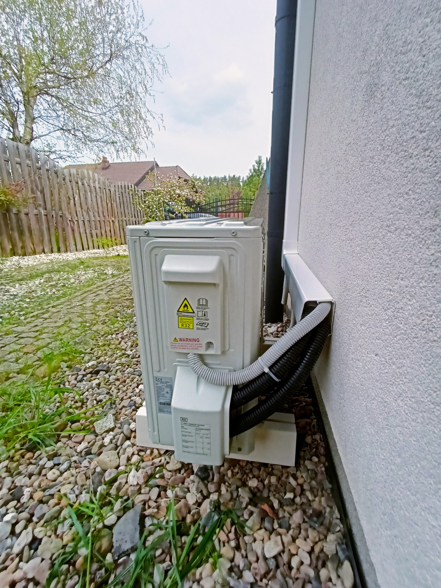 Zewnętrzna jednostka klimatyzacji zamontowana przy ścianie domu, widoczne przewody w osłonie, tło z kamieniami, trawa i drewniany płot.