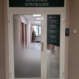 Wejście do kancelarii adwokackiej z informacją o adwokatach i numerami telefonów, widoczne przez szklane drzwi z dekoracyjnym wzorem kropek.