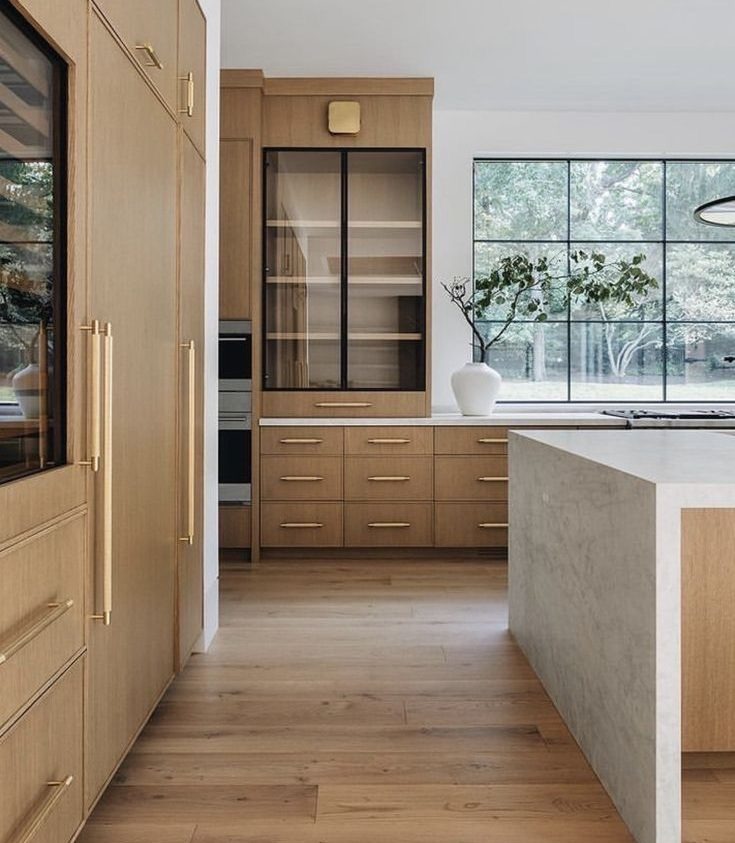 Jasna drewniana kuchnia z wyspą i dużym oknem. Fronty szafek z poziomymi uchwytami. Szklana witryna z czarną ramą. Minimalistyczny design.