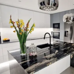 Nowoczesna kuchnia z wyspą o blacie z czarnego kamienia, kwiatami w wazonie i chromowanymi lampami. Białe i szare fronty szafek, sprzęt AGD w zabudowie.