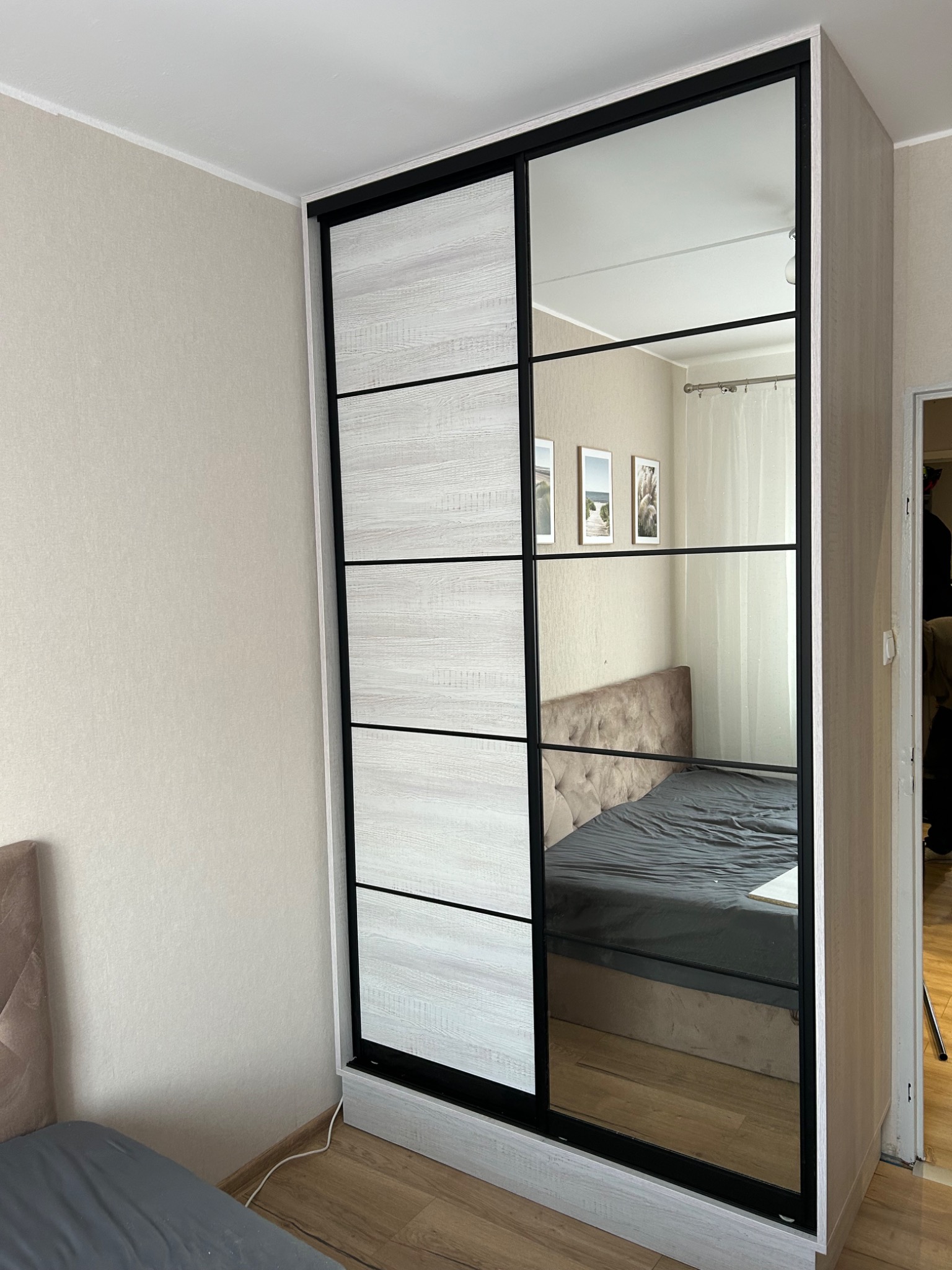 Szafa wnękowa z przesuwnymi drzwiami, część z lustrem, druga z panelami drewnopodobnymi w odcieniach bieli i szarości, czarne ramy, widok na wnętrze pokoju.