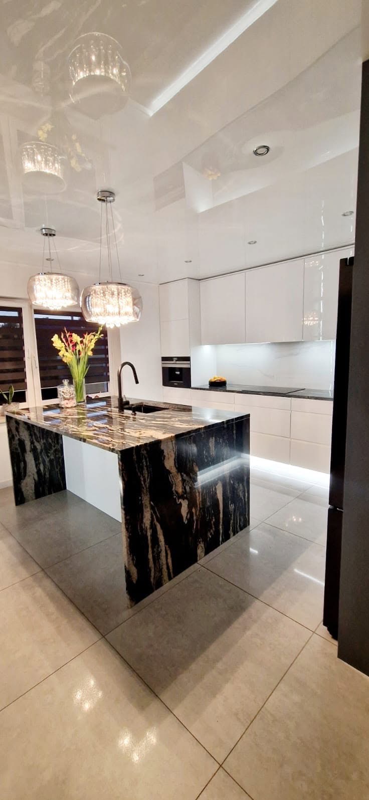 Nowoczesna kuchnia z wyspą o marmurkowym blacie i białymi szafkami. Nad wyspą wiszą ozdobne lampy, a pod szafkami zamontowano oświetlenie LED.