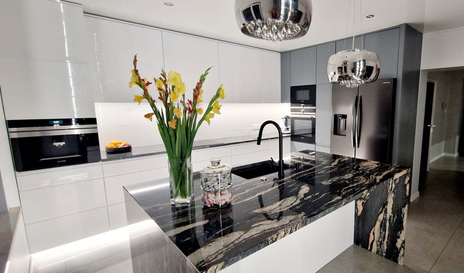 Nowoczesna kuchnia z wyspą o blacie z czarnego kamienia, kwiatami w wazonie i chromowanymi lampami. Białe i szare fronty szafek, sprzęt AGD w zabudowie.