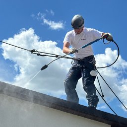 DKMAL MALOWANIE DACHÓW - Pracownik w kasku myje dach budynku za pomocą myjki ciśnieniowej. Widoczne chmury na niebie i lanca myjąca w użyciu.