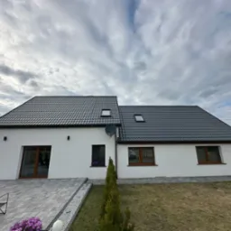 Nowoczesny dom z ciemnoszarym dachem i białymi ścianami, drewniane okna, kostka brukowa, tło: pochmurne niebo. Widoczna antena satelitarna na elewacji.