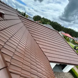 Ciemnobrązowy dach z dachówki ceramicznej, świeżo po renowacji, widok z góry na tle pochmurnego nieba. Widoczne detale tekstury dachówki i otoczenie domu.