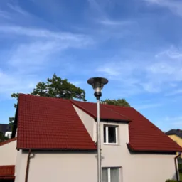 Dom z czerwonym dachem i jasną elewacją w słoneczny dzień. Widoczna lampa uliczna i fragmenty innych budynków w tle. Czyste niebo z delikatnymi chmurami.