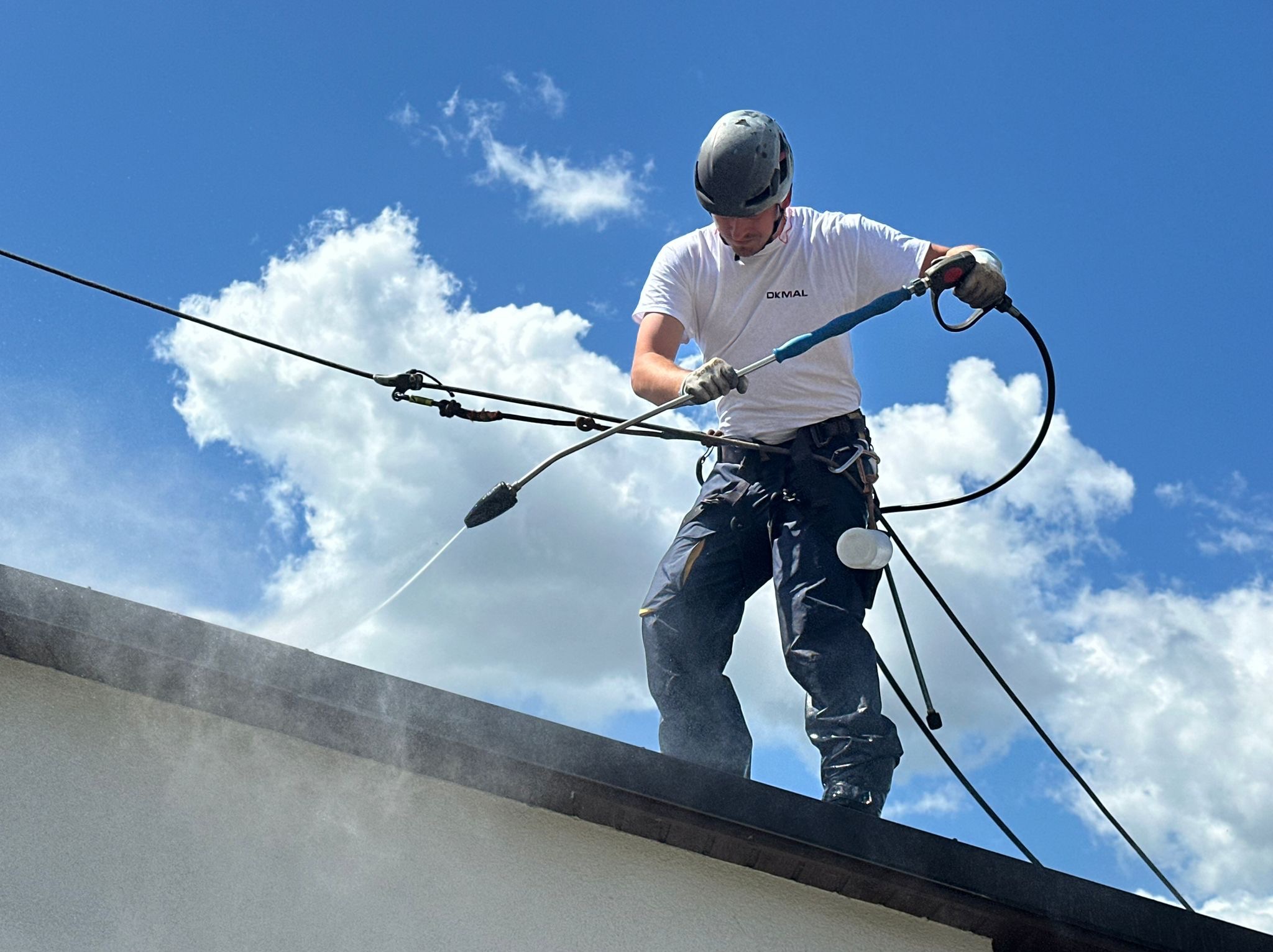 Pracownik w kasku myje dach budynku za pomocą myjki ciśnieniowej. Widoczne chmury na niebie i lanca myjąca w użyciu.