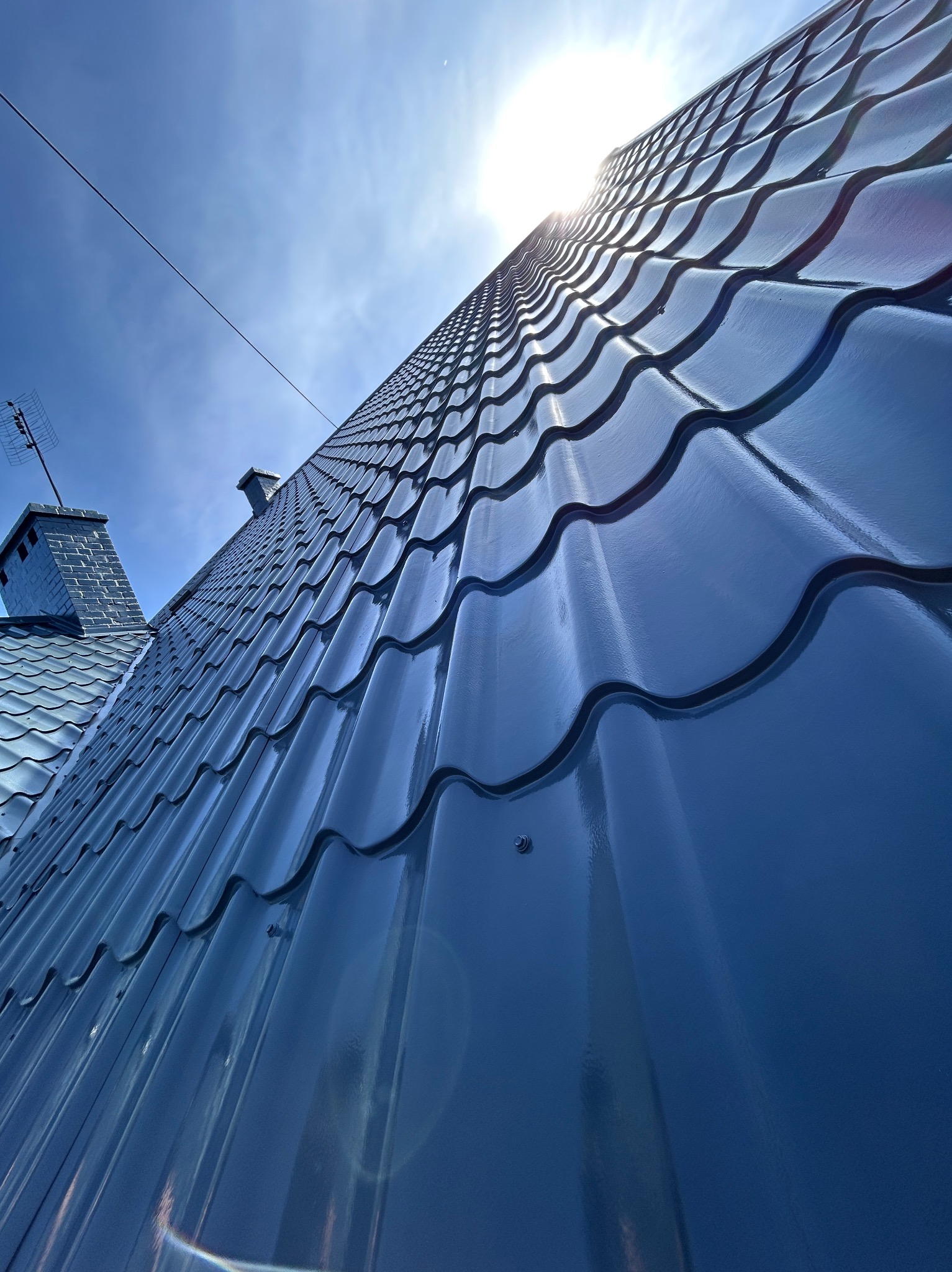 Błyszczący, pomalowany na niebiesko dach z dachówki ceramicznej w Gryficach, uchwycony pod słońce z niskiego kąta, z widocznym kominem i anteną.