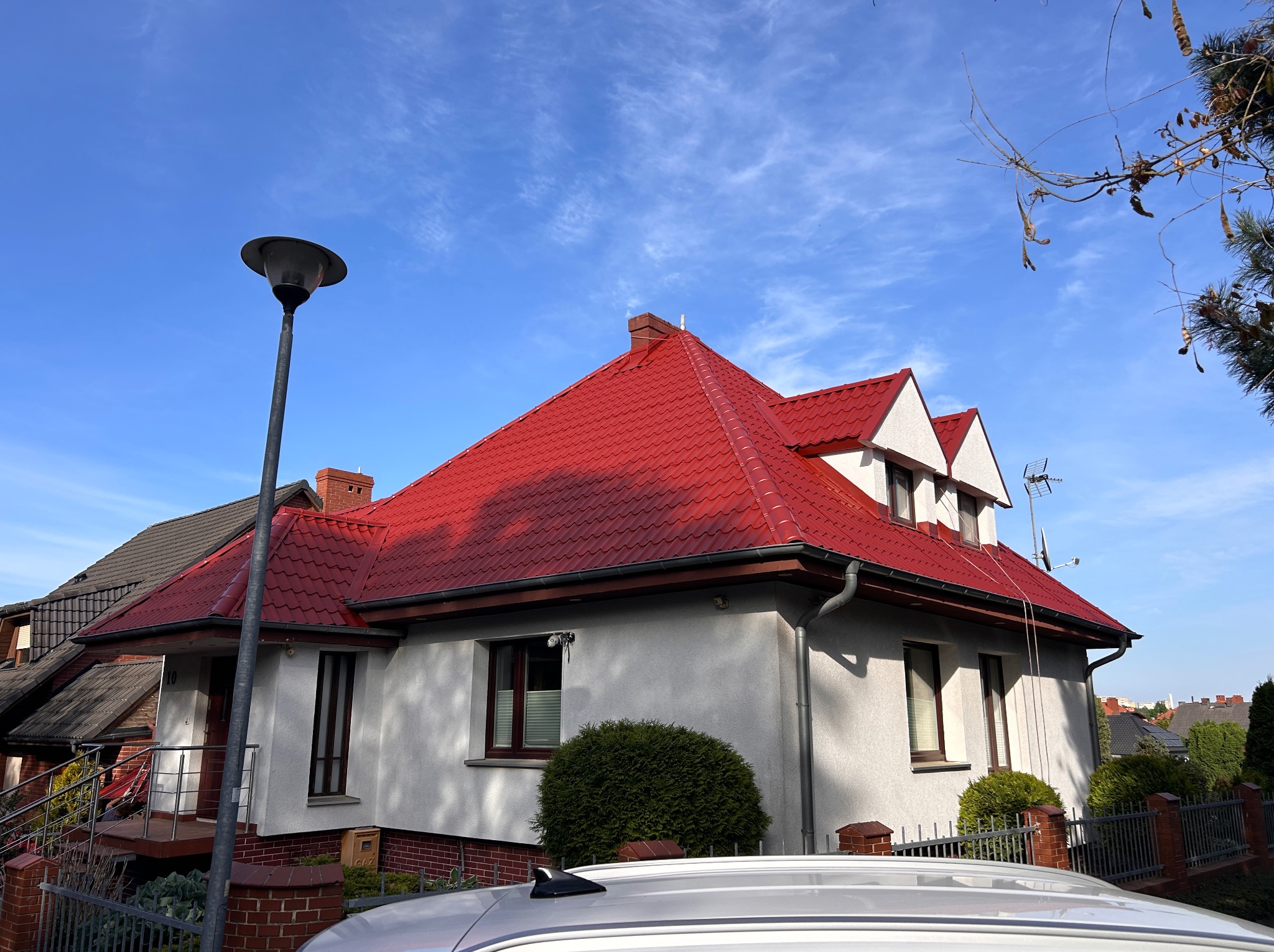 Dom jednorodzinny z czerwonym dachem w blasku słońca, widoczny fragment białego samochodu. Ujęcie z dołu podkreśla geometrię dachu i detale elewacji.