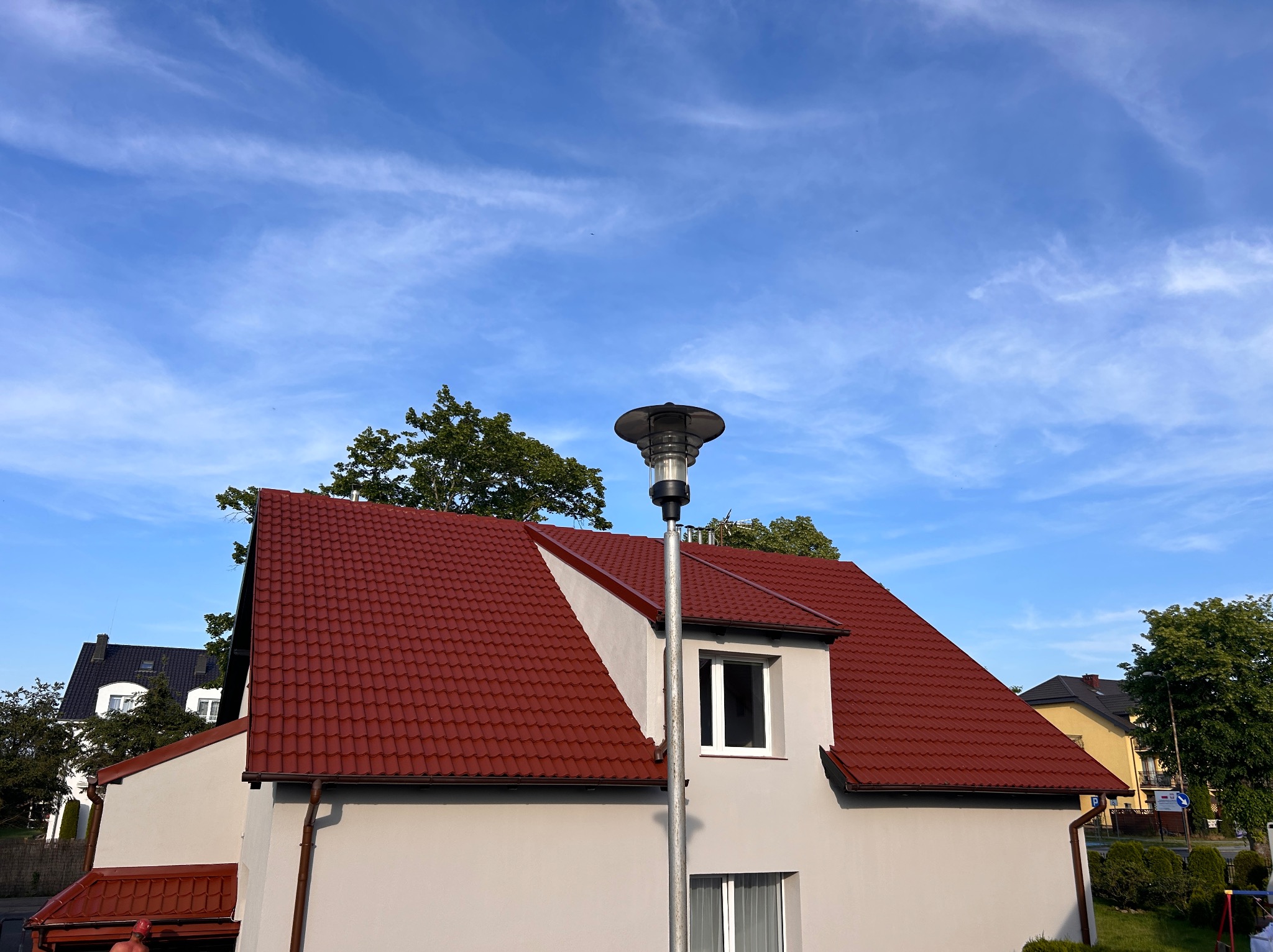 Dom z czerwonym dachem i jasną elewacją w słoneczny dzień. Widoczna lampa uliczna i fragmenty innych budynków w tle. Czyste niebo z delikatnymi chmurami.