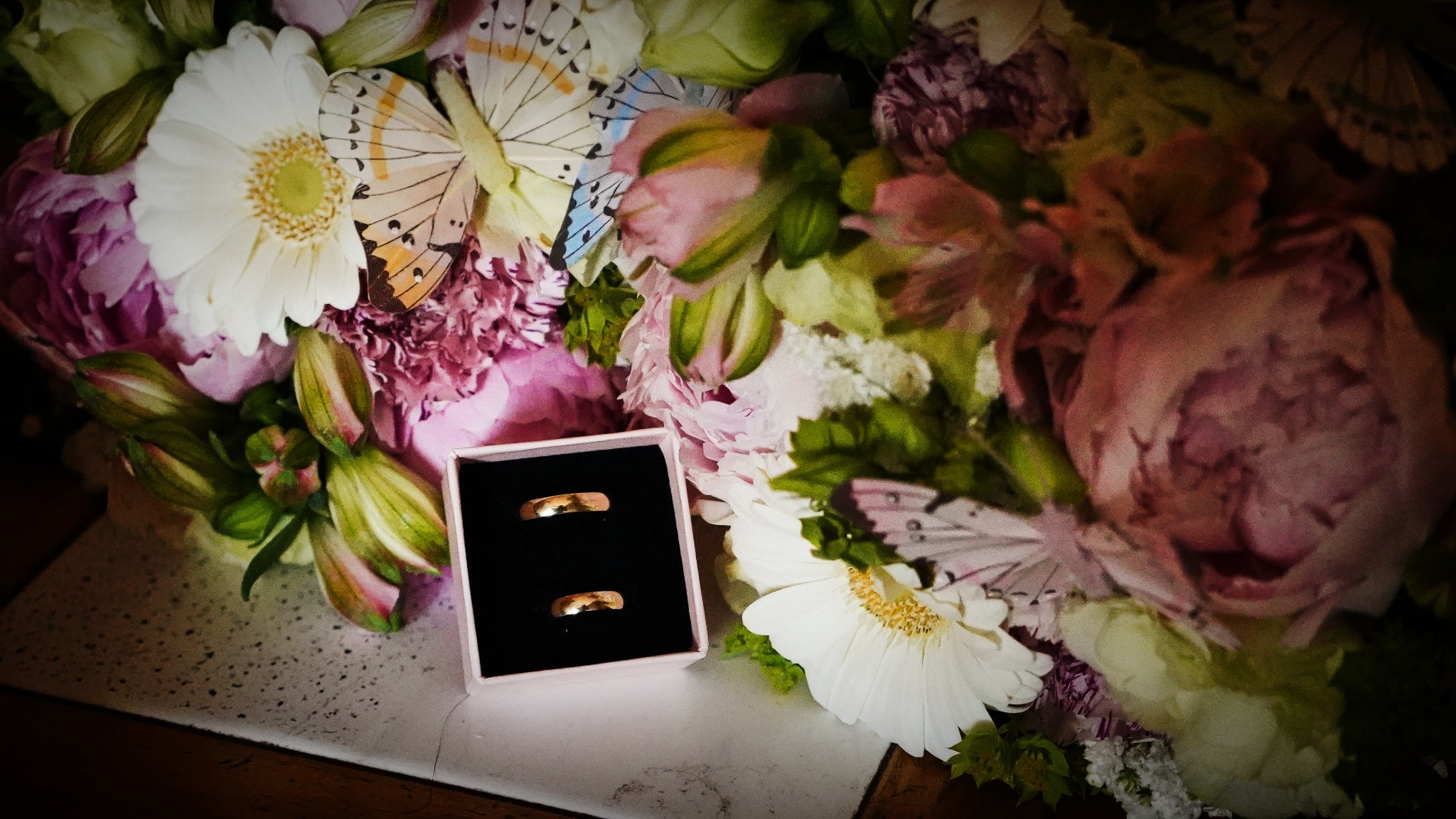Dwa złote pierścionki ślubne w czarnym pudełku na tle bukietu z różowych piwonii, białych gerber i dekoracyjnych motyli.