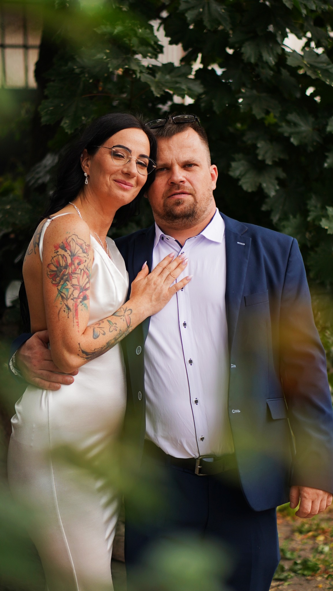 Para w eleganckich strojach pozuje na tle zieleni, kobieta w białej sukience z tatuażem na ramieniu, mężczyzna w granatowej marynarce i białej koszuli.