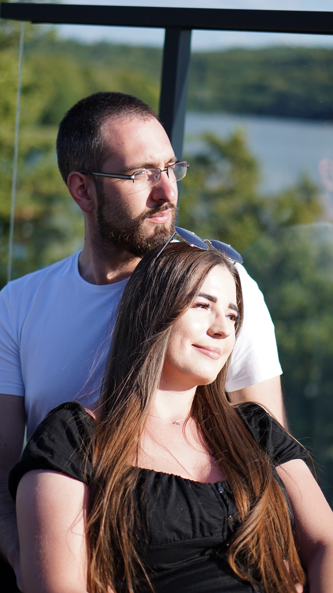 Młoda para, mężczyzna z brodą i okularami obejmuje kobietę z długimi brązowymi włosami i okularami na głowie, na tle zieleni i wody.