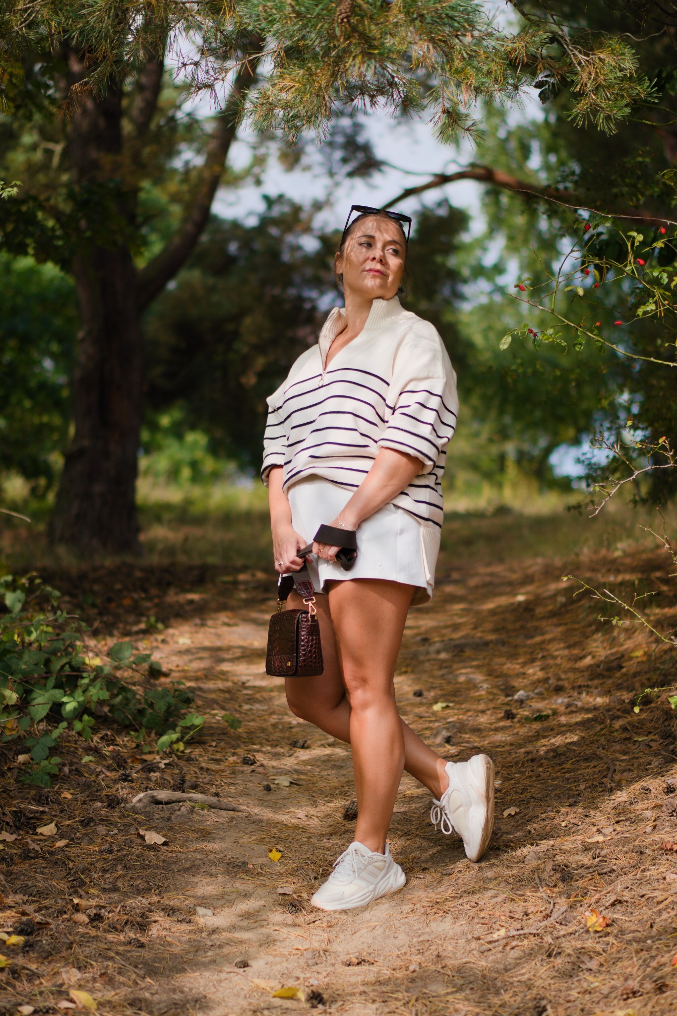 Kobieta w paski w kremowym swetrze i białych szortach pozuje z brązową torebką na leśnej ścieżce, z promieniami słońca padającymi przez korony drzew.