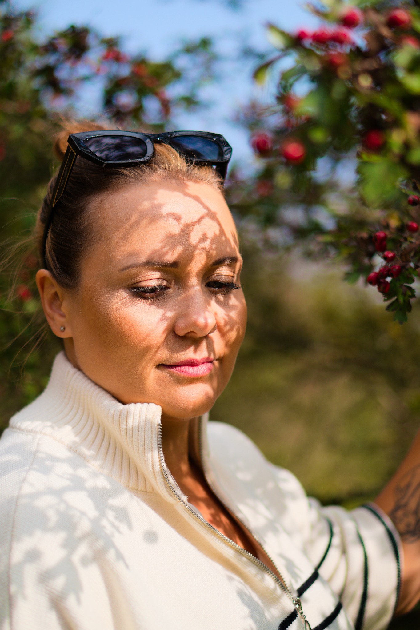 Portret kobiety w kremowym swetrze z suwakiem, z okularami na głowie, w plenerze, z cieniami liści na twarzy i tatuażem na ramieniu.