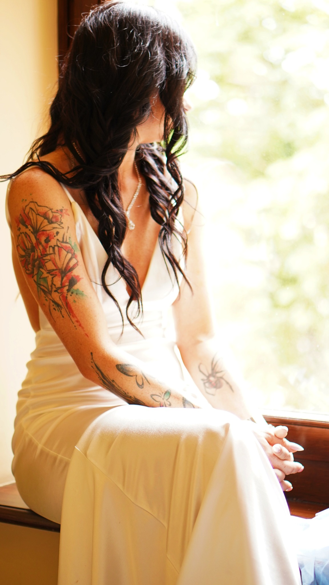 Kobieta w białej sukni siedzi przy oknie, z widocznymi tatuażami na ramionach i dekolcie, spoglądając w dal.